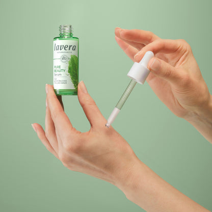 A person holds a small green bottle labeled lavera Pure Beauty Serum while using a white dropper to apply serum to the back of their hand, against a solid sage green background.