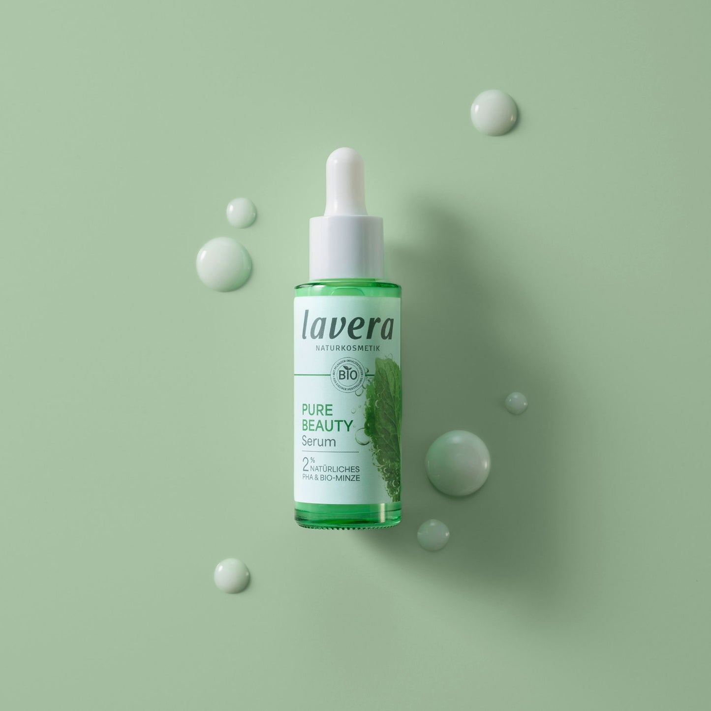 Lavera Pure Beauty Serum in a clear dropper bottle with green liquid, displayed on a light green background with decorative droplet props.