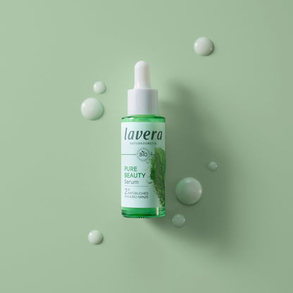 Lavera Pure Beauty Serum in a clear dropper bottle with green liquid, displayed on a light green background with decorative droplet props.