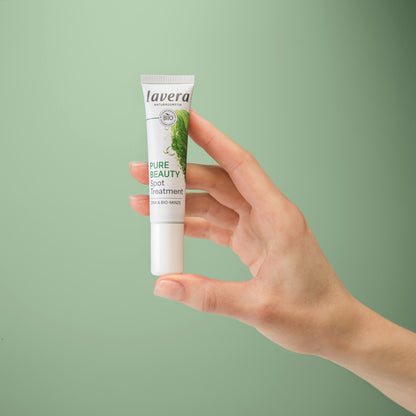 A hand holding a white tube of Lavera Pure Beauty Spot Treatment against a solid sage green background.