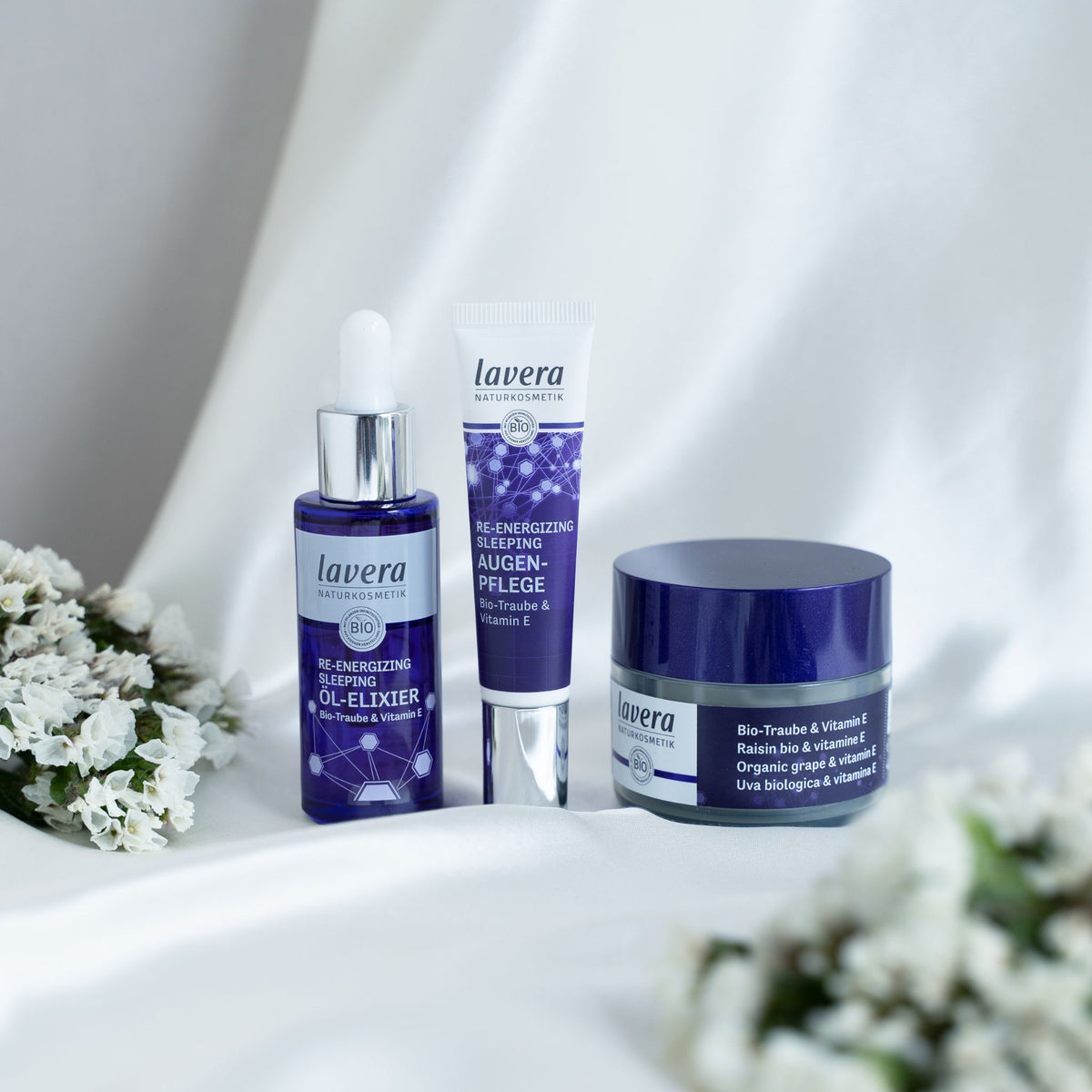 Three Lavera Natural skincare products arranged on draped white fabric with small white flowers: a purple dropper bottle labeled Re-Energizing Oil-Elixir, a purple tube labeled Re-Energizing Eye Cream, and a dark blue jar labeled Re-Energizing Sleeping Cream.