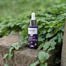 A bottle of Lavera Organic Re-Energizing Natural Oil-Elixir, featuring a dark purple bottle and white dropper cap, resting on a stone ledge surrounded by lush green foliage.