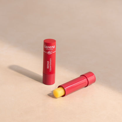 Lavera lip balm stick in a red tube standing upright, with the second tube lying horizontally and its yellow balm stick exposed, on a warm beige background.