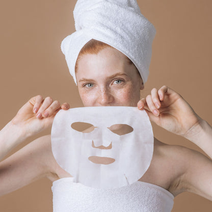 A woman with a white towel wrapped around her head holds a white sheet mask in front of her face against a solid beige background.
