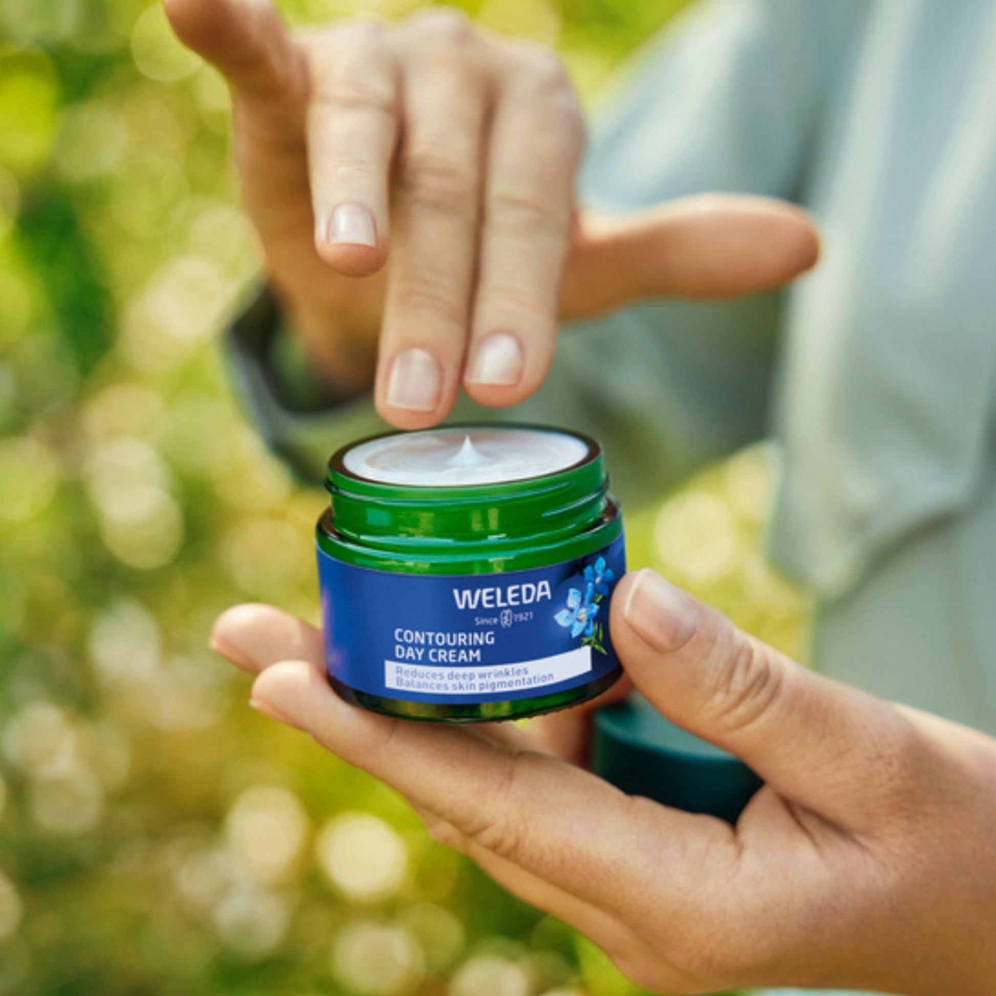 A person outdoors holds an open jar of Weleda Contouring Day Cream, with the cream visible on the lid and a blurred green garden background.