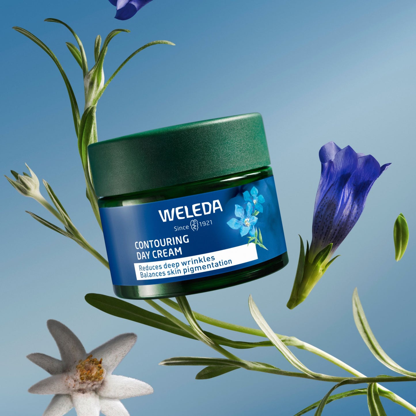 A jar of Weleda Contouring Day Cream floating against a blue sky background with botanical elements including a blue flower, a white flower, and green leaves. The jar is dark green with a blue label.