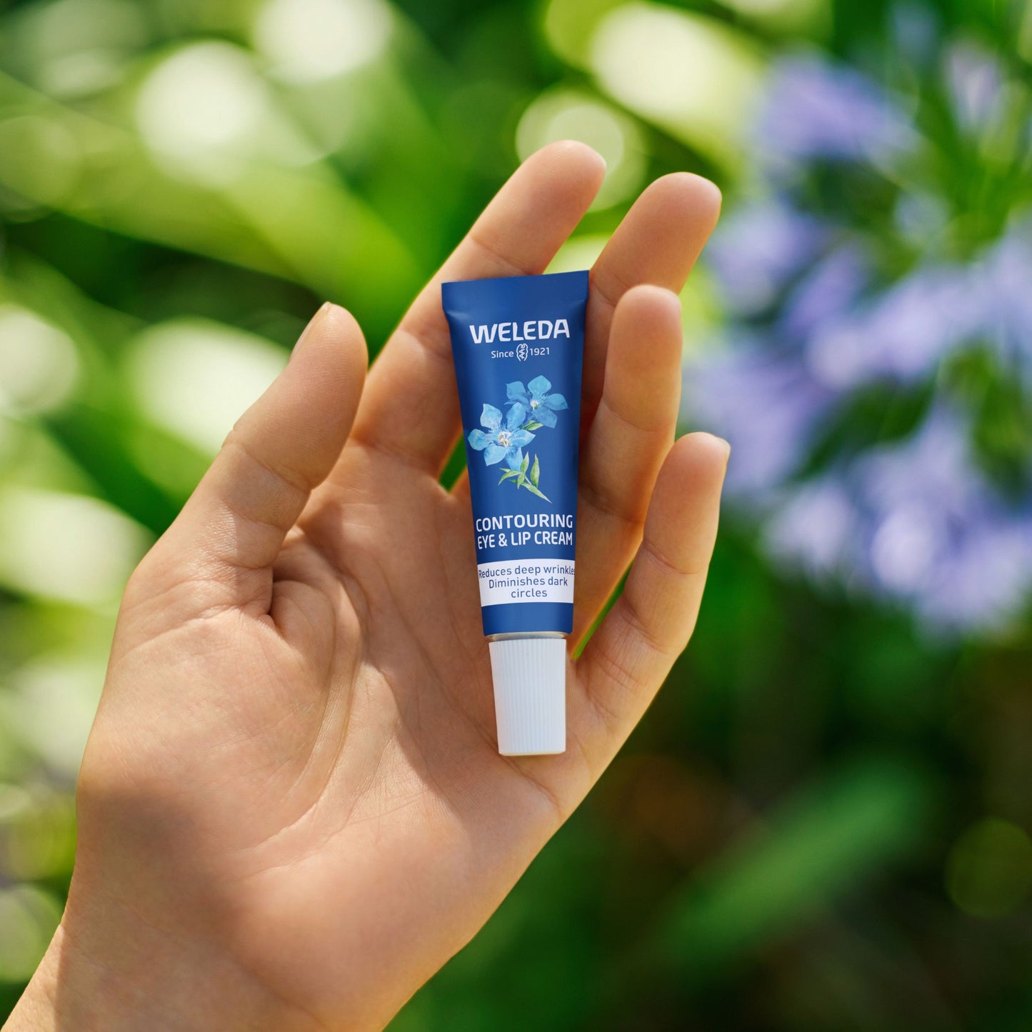 A tube of Weleda Contouring Eye & Lip Cream on a hand with natural green and purple flower background. The tube is dark blue with white text and a graphic of blue forget-me-not flowers, with a white ribbed cap.