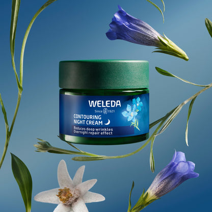 A jar of Weleda Contouring Night Cream floating against a blue gradient background, surrounded by botanical elements including blue bellflowers, green leaves, and a white flower. The product is a dark green jar with a blue label featuring white text and a small blue flower graphic.