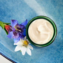 A jar of Weleda Contouring Night Cream against a blue gradient background, surrounded by botanical elements including blue bellflowers and a white flower.