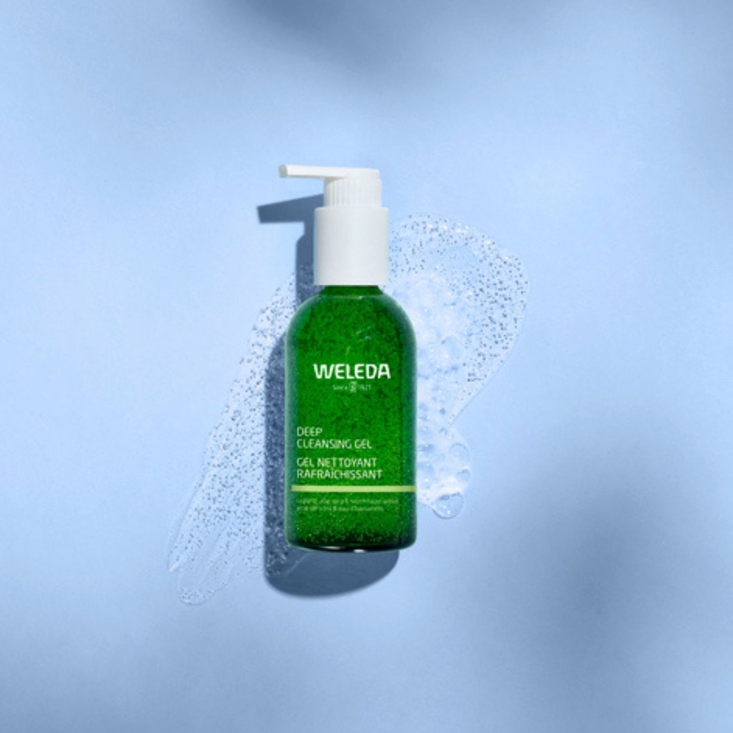 A green bottle of Weleda Deep Cleansing Gel with a white pump dispenser, set against a light blue background with a splash of clear foam around it.