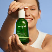 A woman holding a green Weleda Deep Cleansing Gel bottle against a solid blue background.