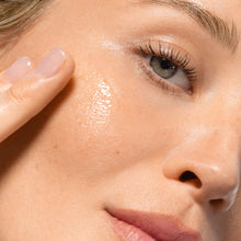 Close-up of a woman's face with glossy, hydrated skin, showing her cheek, eye, nose, and lips. A finger is touching her cheek, highlighting a dewy, radiant complexion.