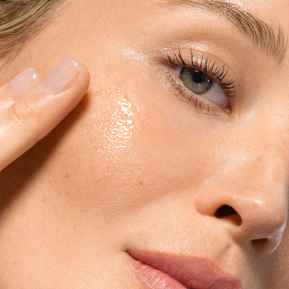 Close-up of a woman's face with glossy, hydrated skin, showing her cheek, eye, nose, and lips. A finger is touching her cheek, highlighting a dewy, radiant complexion.