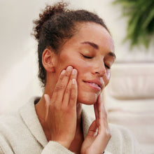 A close-up lifestyle shot of a woman with closed eyes, gently touching her face with both hands, wearing a light beige knit robe. The background is a soft, out-of-focus indoor setting with a hint of green foliage.