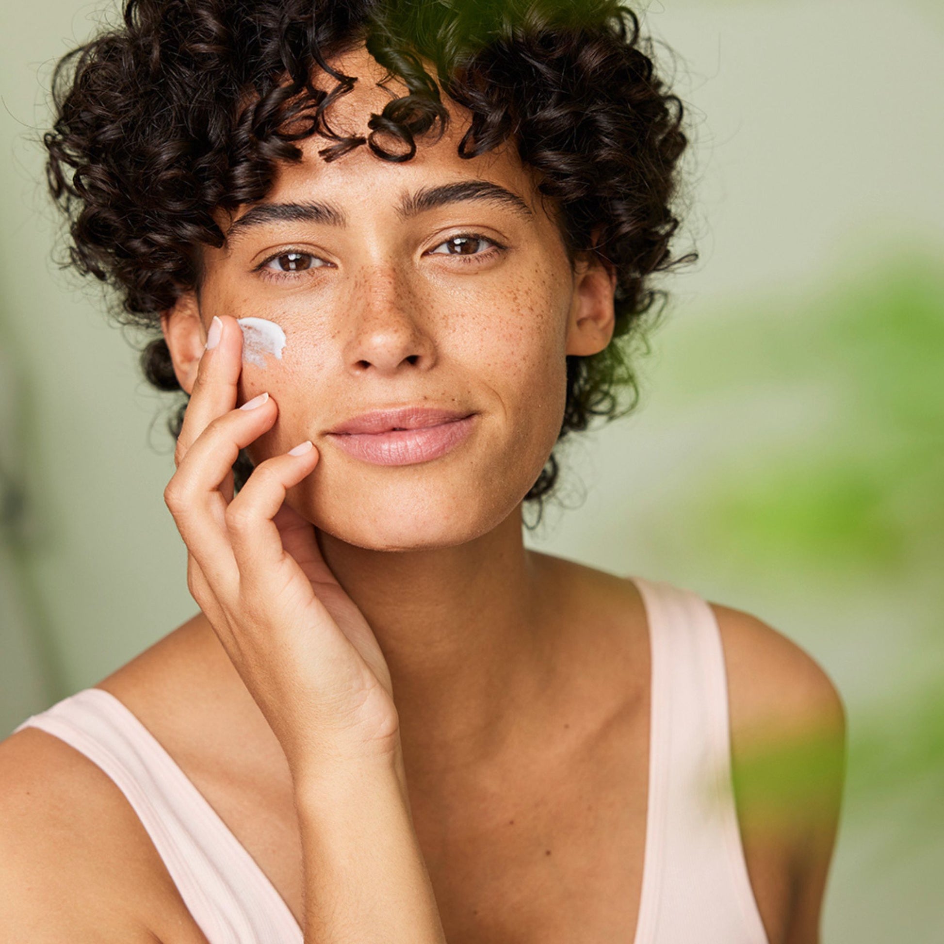 A close-up lifestyle shot of a woman with curly dark hair applying a weleda sensitive soothing moisturising cream to her cheek. She is wearing a light pink tank top against a soft, blurred green background.