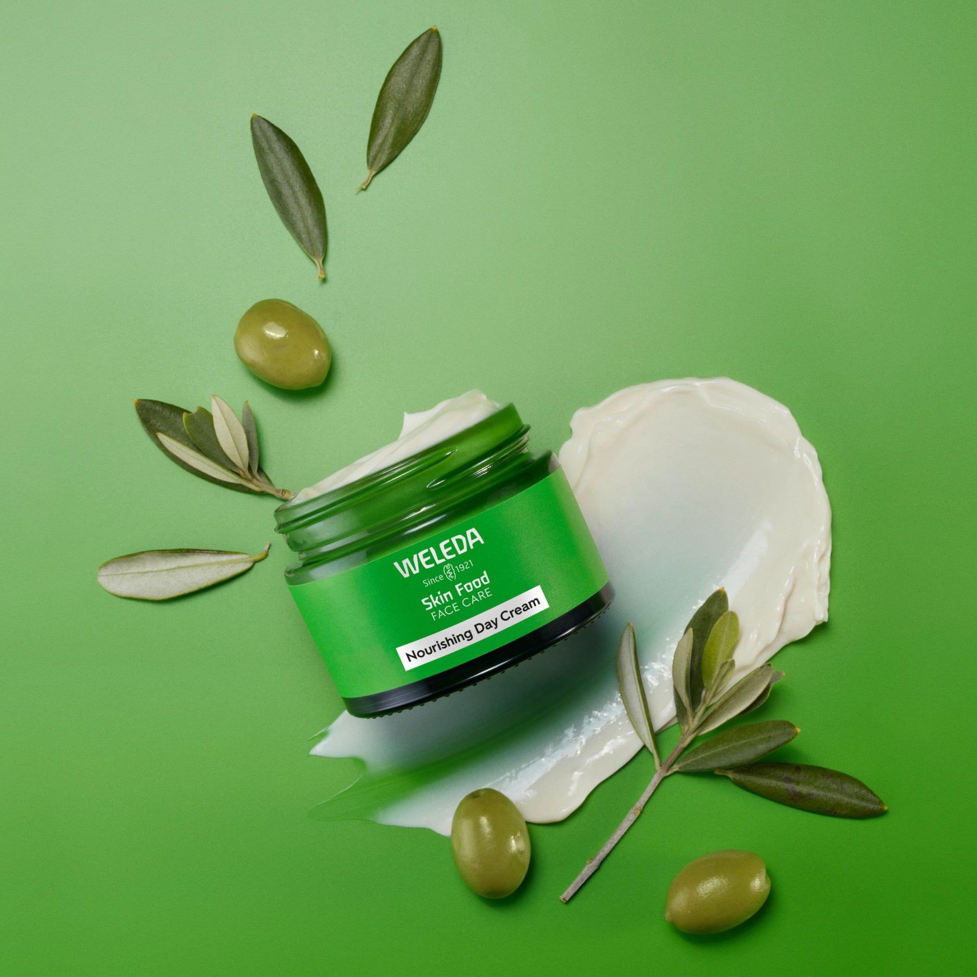 A jar of Weleda Skin Food Face Cream placed on a bright green background with olive branches, olives, and a smear of white cream around it.