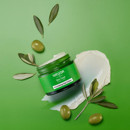 A jar of Weleda Skin Food Face Cream placed on a bright green background with olive branches, olives, and a smear of white cream around it.