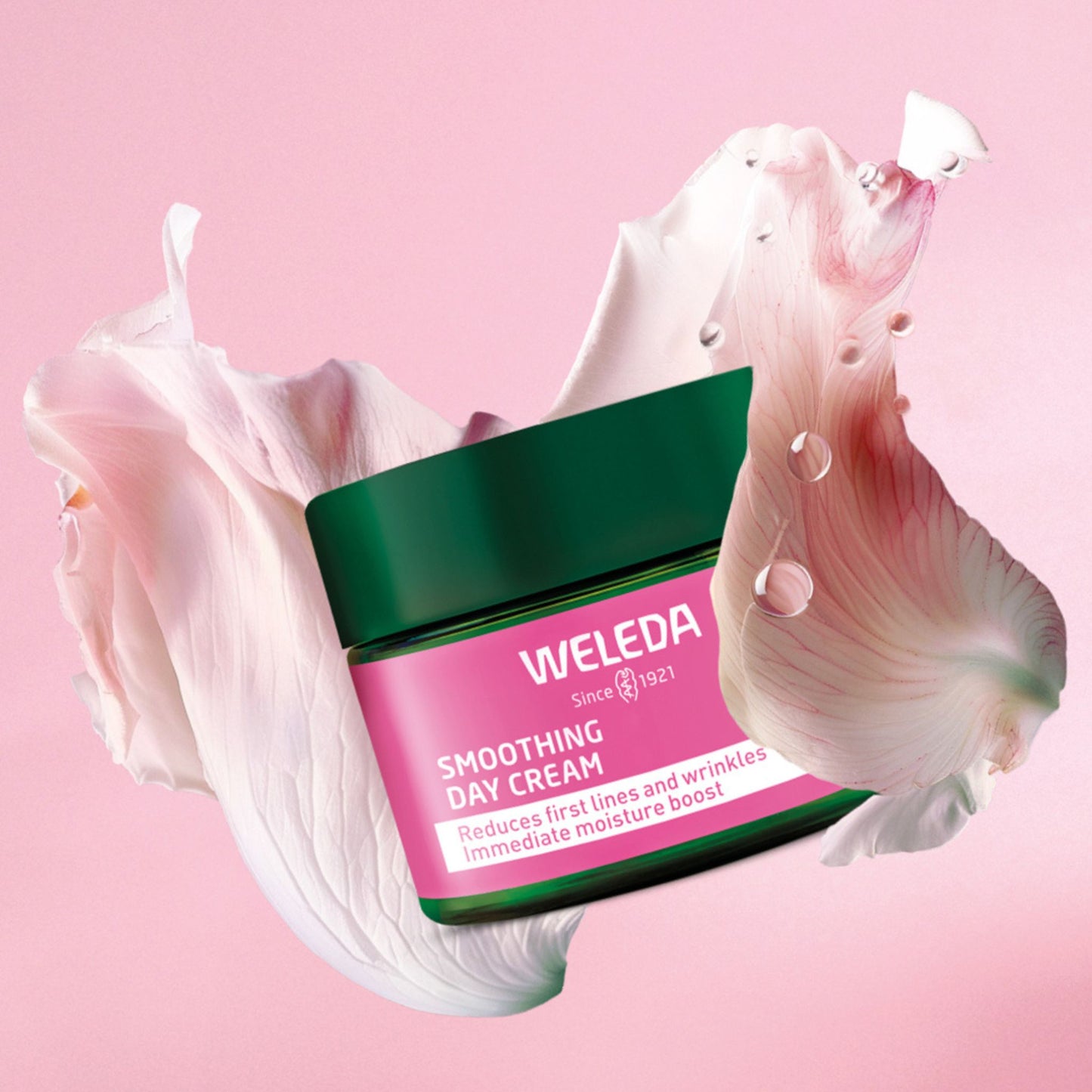 A jar of Weleda Smoothing Day Cream centered against a soft pink background, surrounded by rose petals and water droplets. The jar has a dark green lid and a pink label with white text.