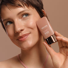 A close-up lifestyle shot of a woman with short brown hair holding a bottle of Weleda Sun-kissed Bronzing Serum Drops next to her cheek, set against a solid warm beige background.