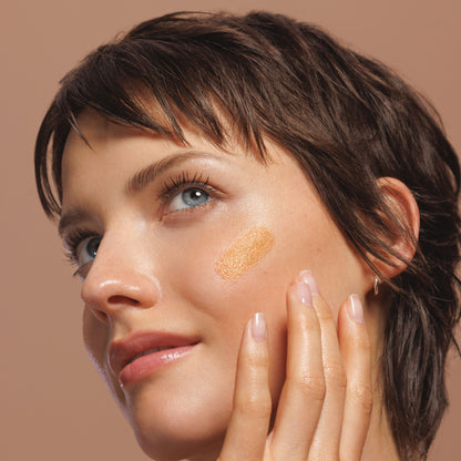 Close-up of a woman with short dark brown hair applying or showing a sun-kissed serum booster liquid swatch on her cheek against a solid warm beige background.